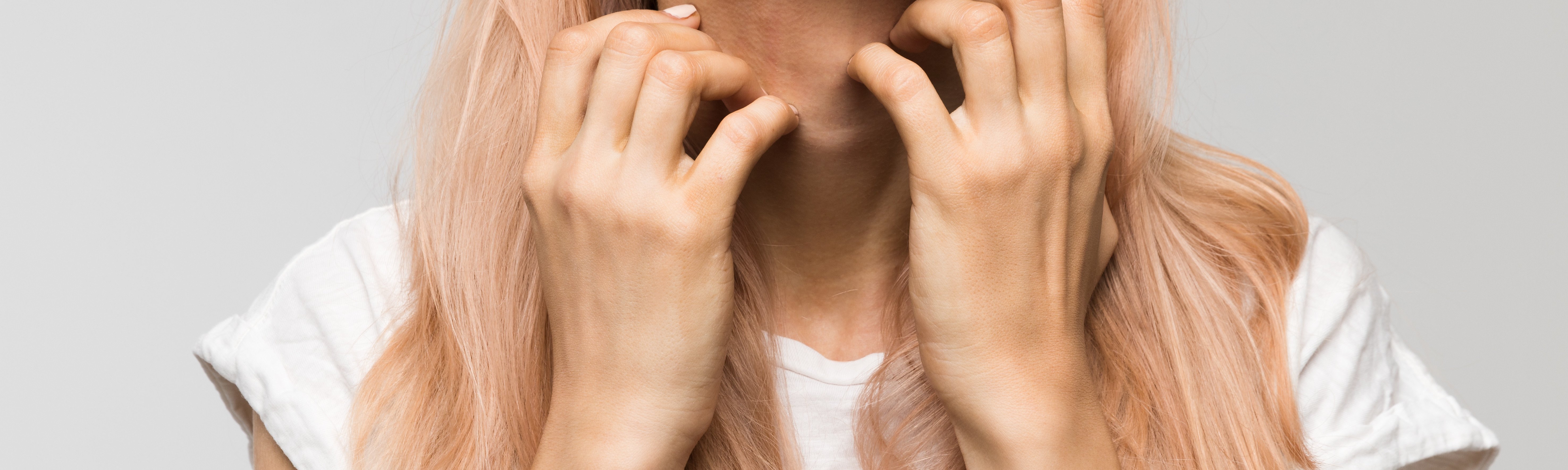 woman with light pink hair scratching with both hands at the skin on her neck wearing a white tee shirt on a gray background